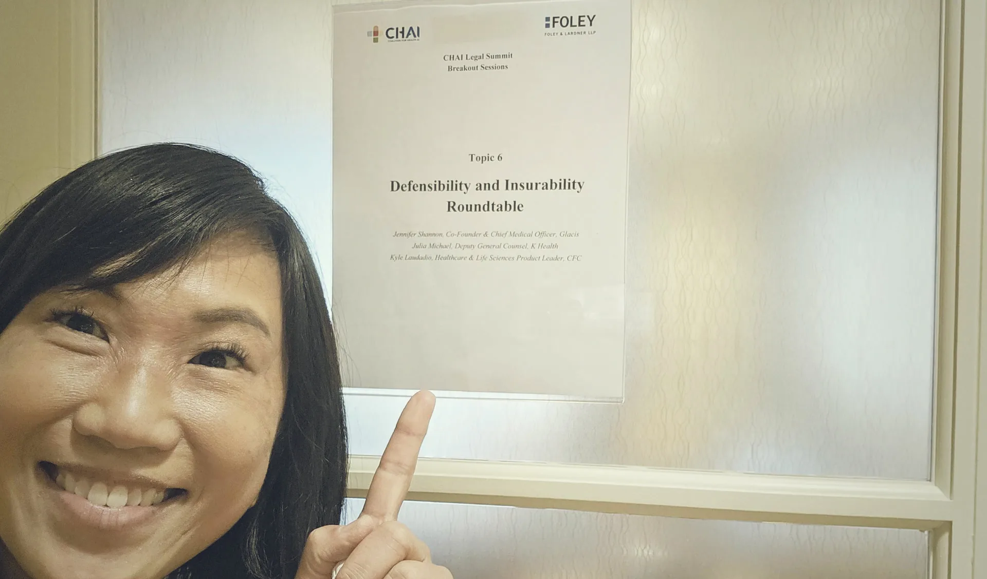 Jennifer Shannon pointing at the printed sign for Topic 6, the Defensibility and Insurability Roundtable at the CHAI Legal Summit, which lists the three speakers.