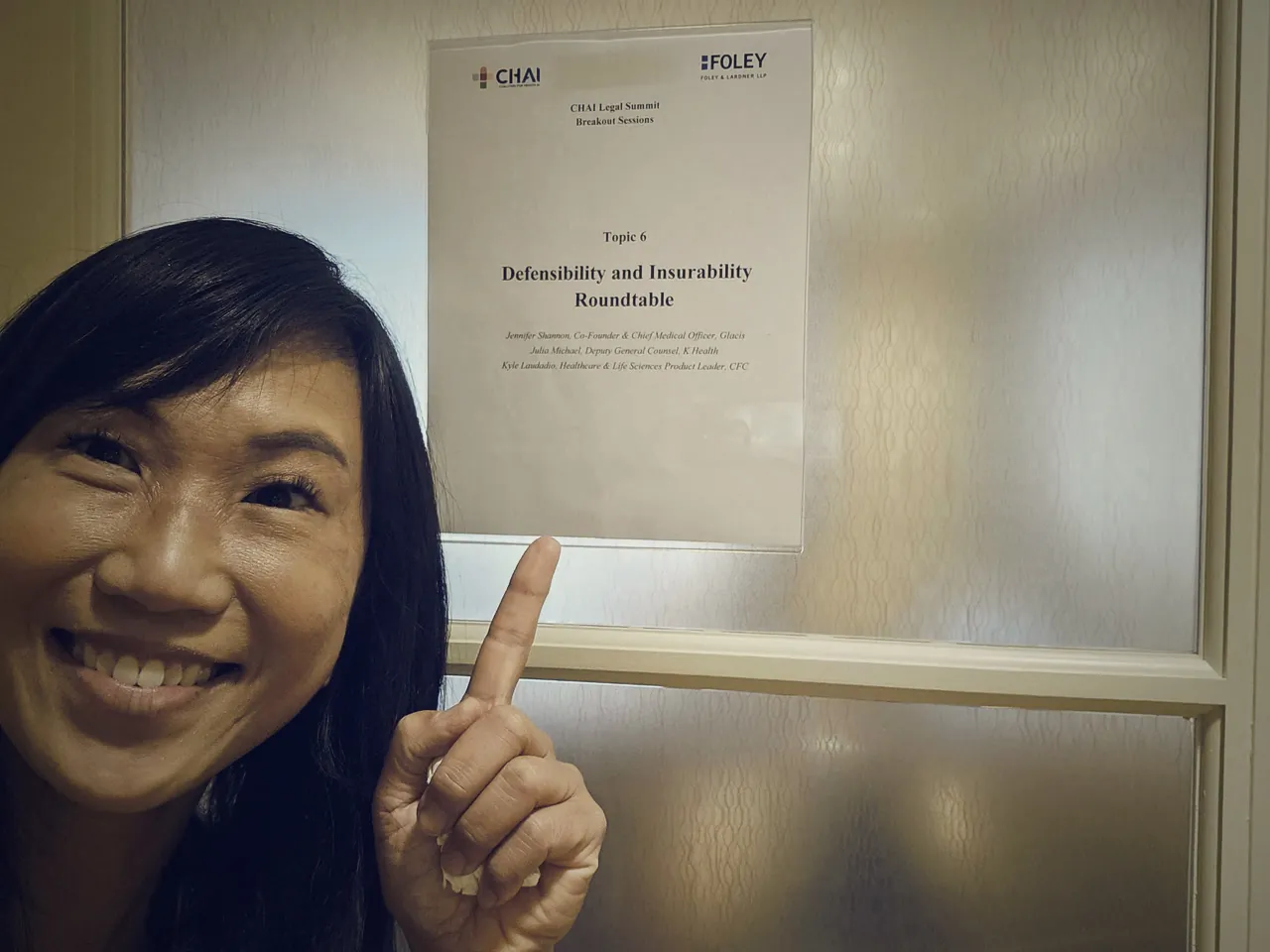 Jennifer Shannon pointing at a printed sign for Topic 6, the Defensibility and Insurability Roundtable at the CHAI Legal Summit, listing the three speakers.